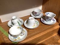 Five porcelain tea cups and saucers arranged on wooden shelf, showing general condition and floral patterns.