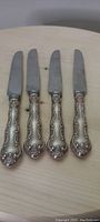 Four Birks Chantilly lace pattern sterling silver handled dinner knives laid out on light wood surface, showing the full length of the knives.