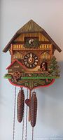 Front view of the colorful wooden cuckoo clock depicting a chalet with a clock face, two pinecone-shaped weights, and decorative figurines.