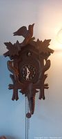 Front view of wooden brown cuckoo clock showing carved leaves, bird on top, clock face and pinecone weights.
