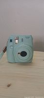 Front view of light blue Fujifilm Instax Mini 9 camera showing lens, flash, shutter button, and selfie mirror.