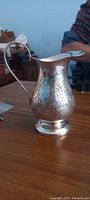 Silver creamer jug with floral engraving viewed from the side on a wooden table, showing handle and spout.
