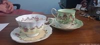 Photo of two Royal Albert bone china tea cups and saucers on a wooden surface, one yellow floral Invitation Series set and one green botanical Jack-in-a-Pulpit set