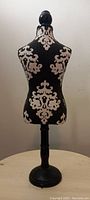 Frontal view of small fabric-covered black and beige patterned mannequin necklace display on round black pedestal stand.