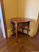 Single circular wooden side table positioned in a corner on hardwood floor, showing legs and open slat perimeter below tabletop.