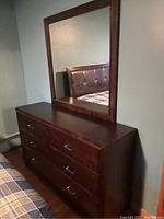 Front-left angled view of a rectangular wooden dresser with six drawers and metal handles, topped with a large square mirror with a dark wood frame, placed in a bedroom next to a bed.