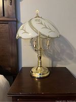 Brass tavern style table lamp with white glass shade and gold decoration shown on wooden side table.