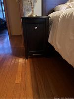 Front view of black wood night stand next to bed and on wooden floor, showing silver knob on door and flat top.