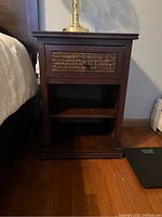 Front view of wooden night stand showing drawer with woven front panel and open shelf below.