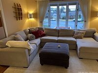 Angled view of a light gray fabric sectional sofa with several cushions and pillows in various colors. Sofa positioned against two walls with window and lamp visible.
