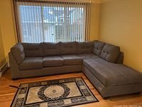 Photo shows a large grey sectional sofa placed against a window with vertical blinds, displaying the full layout with chaise lounge on the right side.