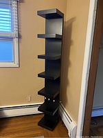 Tall black wood corner shelving unit shown standing in a room corner next to a window and baseboard heater, illustrating its height and design.