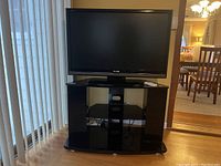 Front view of TV stand with flat screen TV placed on top. TV stand has black glass shelves and black metal supports.