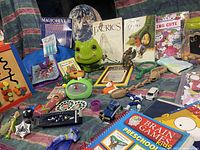 Photo showing an assortment of kids items including toys, books, and games spread over a colorful fabric background. Items visible include plush frog toy, wooden bead maze, Magic Eye II book, various small toys, and educational card game.