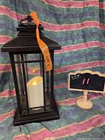 Front and side view of black metal carriage lantern with a lit battery-powered candle inside, showing the design and condition including the damaged hinge.