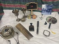 Curio lot items arranged on white cloth with patterned background including silver goblets, colorful Thai compacts, vintage fan, hand-carved combs, glass bottle, silver-tone picture frame, and decorative pendant.