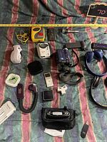 Photo shows assortment of vintage electronics on a striped fabric background including 2 Sony Walkmans, a blue Sony digital video camera, black compact camera, headphones, iPods, and accessories.