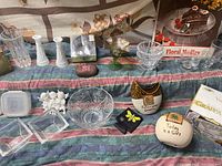 Photo displaying a variety of glassware and decor items including bowls, vases, figurines, and a cake platter arranged on a table covered in fabric.