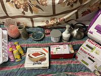 Full view of the lot including Starfrit spiralizer boxes, Pyrex baking dish box, ceramic vases, and kitchen tools laid out on fabric.