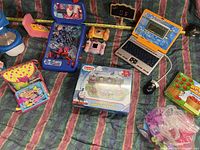 Overview of all items in lot arranged on a fabric background, showing multiple toys including educational laptop, puzzle tin, doll set, magnifier