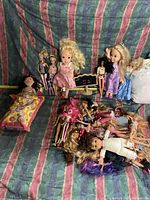 Photo showing multiple Barbie and fashion dolls standing and lying on a striped fabric background, with doll bed and accessories visible