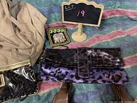 Photo showing brown and beige cloth handbag, shiny purple-black leopard print clutch, small black mesh/net bag with gold chain strap all laid on colorful fabric.