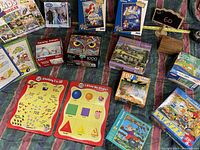 Image shows assortment of multiple jigsaw puzzle boxes of various sizes and themes including Disney, animals, landscapes, and educational puzzles spread out on cloth surface.