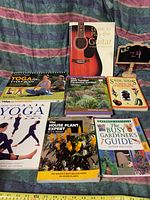 Photo of seven self improvement books laid out on a cloth background with a ruler showing scale. Titles related to yoga, gardening, seed saving, and guitar instruction.