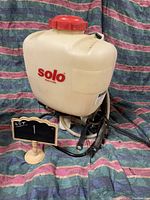 Front view of the white plastic Solo pressurized sprayer tank with red cap and black shoulder straps on a fabric background.