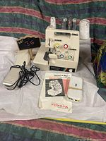 Front and side view of Toyota 14 thread serger sewing machine with instruction manual, foot pedal, and accessory box on white fabric background.