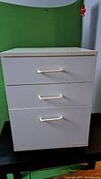 Light beige metal 3 drawer file cabinet on black table against green background