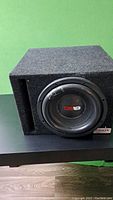 Black DS18 subwoofer speaker in carpeted enclosure with front slot port, sitting on top of black Bbox amplifier, in room with green wall and wood floor.