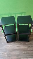 Photo of two black wood end tables, each with a square top and a lower shelf, four legs, on a brown wood floor against a green background.