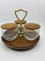 Front view showing the round wooden tray with three glass containers topped with wooden lids and central brass handle.