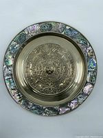 Front view of round metal wall plaque showing embossed Aztec calendar and iridescent seashell border