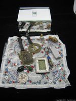 Thirteen miscellaneous vintage trinket and decor items laid out on a white handkerchief with floral and fan patterns, alongside a glass mirror box in the background. Items include badges, keychains, lighter, picture frame, small bell and other collectibles.