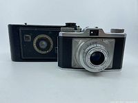 Two vintage cameras side by side, the Braun Nurnberg Gloriette in black with leather case and the Kodak Jiffy collapsible camera