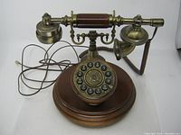 Front view showing wooden base, rotary dial with black number buttons and decorative brass metal detailing, handset cradle, and attached cord.