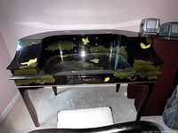 Front view of black lacquer Asian Chinoiserie style writing desk showing gold and green hand-painted motifs and glass top surface.