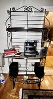 Black metal baker rack with three shelves. Middle shelf holds a stereo system with radio, cassette, and CD capabilities, remote control and two speakers on bottom shelf. Rack has decorative scrollwork.