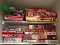 Shelf with assorted boxed board games and kits including Barbie Trace 'n' Color Drawing Set, Snack 'n Go, Mickey Mouse puzzle, Jr. Art Workshop, Ring Toss, Little Pet Pet House, Monkey Business, Bonkers!, Summerfarn, and Monopoly.