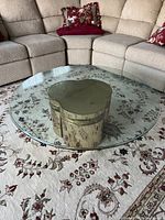 Clear view of the coffee table showing the round glass top and gold pedestal base with floral embossed detail, placed on a patterned rug with beige sectional sofa in background