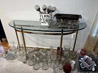 Glass top console table with brass legs viewed from front with various glassware items on top and beneath it including clear and colored glass vases and bowls.