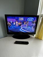Dynex flat screen TV powered on with TV show visible, black remote control in front.
