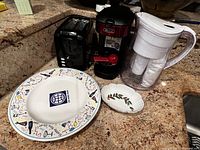Photo showing kitchen counter with water pitcher, coffee maker, toaster, large colorful plate and small oval dish.