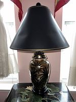 Full view of lamp showing black tapered lampshade and ceramic base with gold floral design on black background.