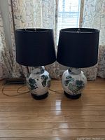 Pair of oriental table lamps with patterned ceramic bases and black drum lampshades sitting on the floor in front of window with floral curtains.