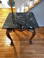 Square black marble top table with carved wooden legs and claw feet, captured indoors on hardwood flooring near stairs.