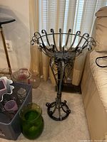 Metal planter stand placed indoors on carpet near window blinds and sofa, showing decorative scrollwork and basket-shaped plant holder