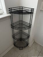Black metal corner shelf unit placed in a corner against white walls and tiled floor, showing the full four-tier wire shelf design.